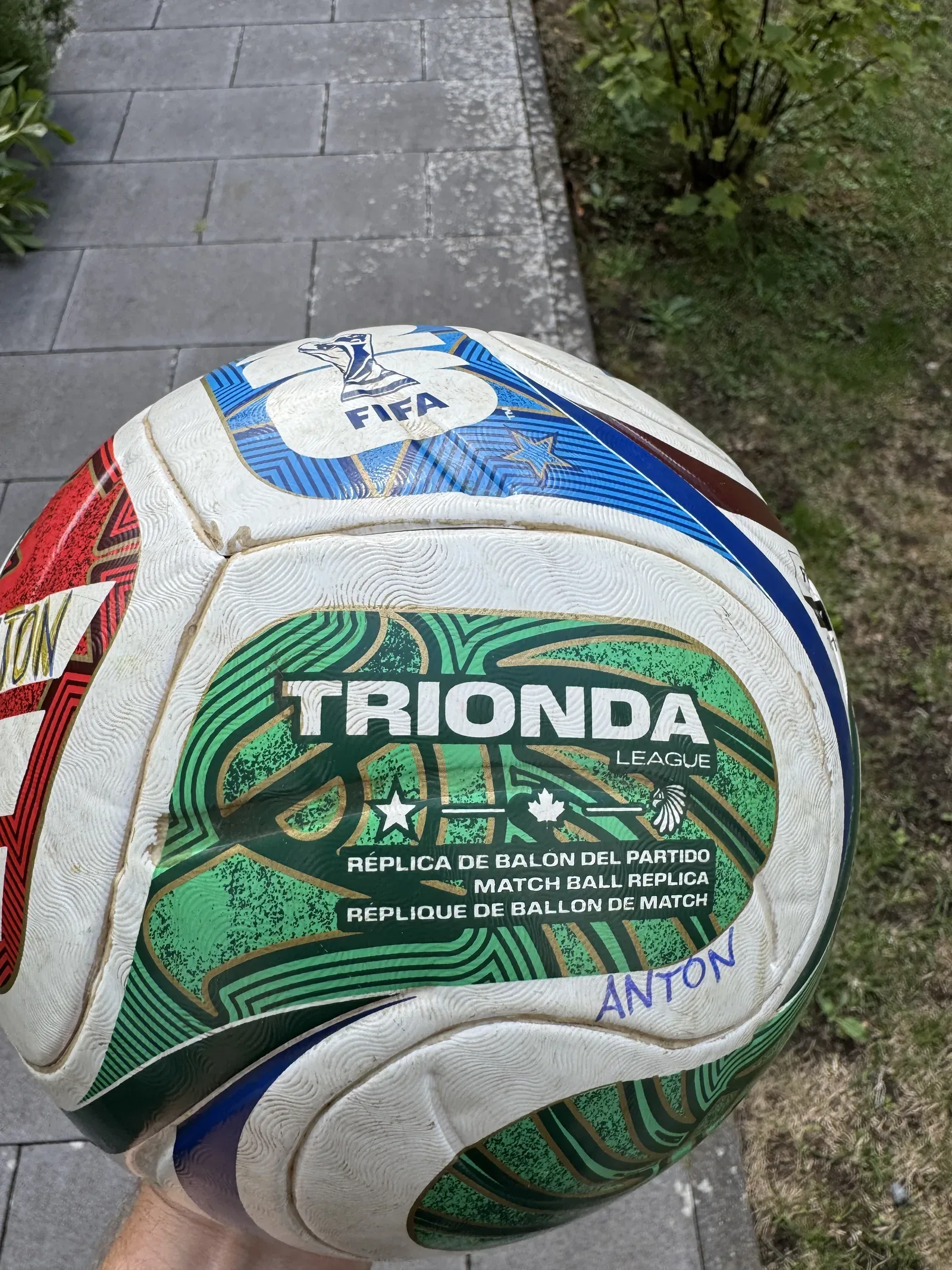 Weißer Fußball mit grünen, roten und blauen Bahnen, in einer Hand gehalten. Lesbar sind das FIFA-Zeichen mit Pokal, der Schriftzug TRIONDA LEAGUE und die Zeile MATCH BALL REPLICA