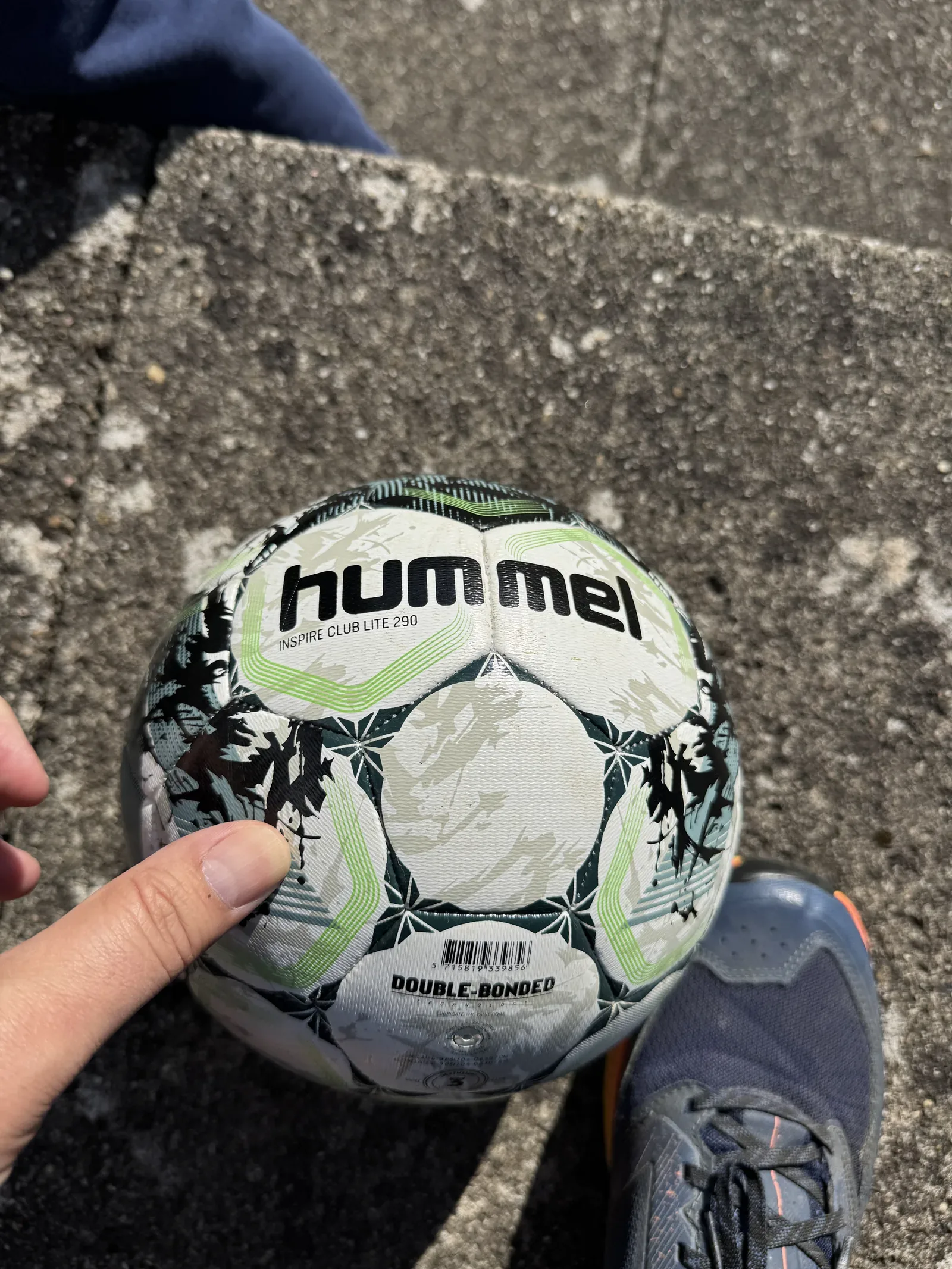Weißer Fußball mit dunkelgrün-hellgrünem Muster in einer Hand über Pflastersteinen. Lesbar sind hummel und darunter INSPIRE CLUB LITE 290.